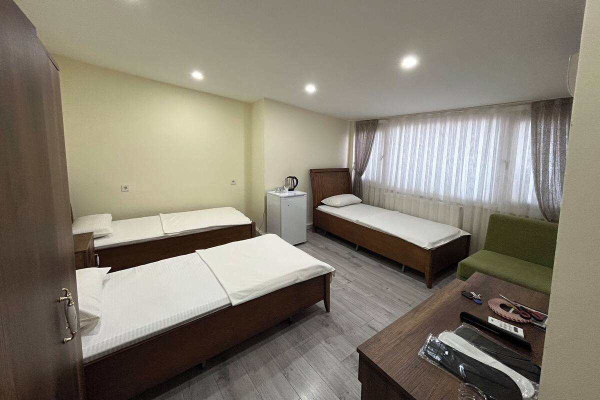 Habitación de hotel con tres camas individuales, una mesita, una silla verde y una ventana con cortinas.