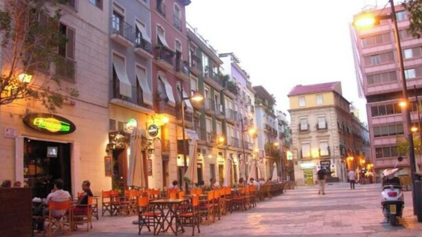 A street with outdoor seating at cafes, buildings with balconies, and a scooter parked on the sidewalk.