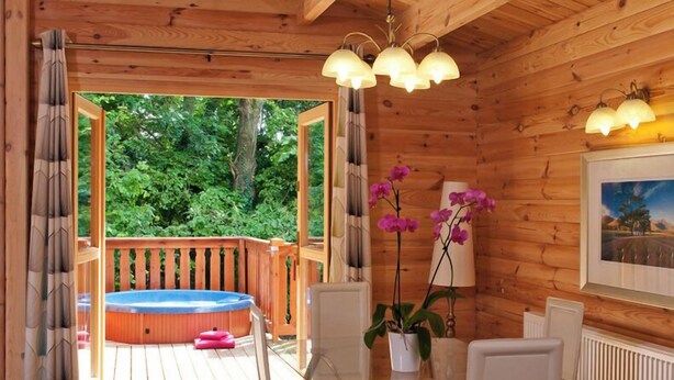 A wooden cabin with a glass table and chairs, a hot tub, and a view of greenery.
