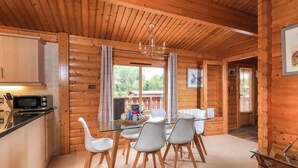 A wooden cabin interior with a dining area featuring a glass table and white chairs, a kitchen with a microwave, and a large window with curtains.
