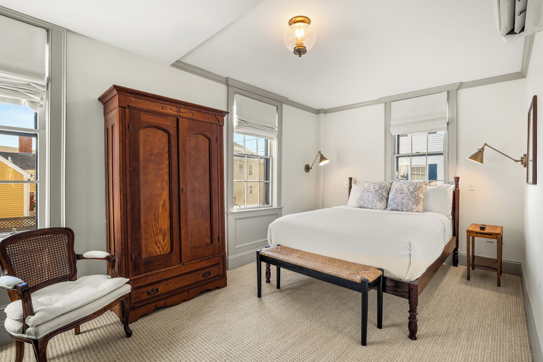 A bedroom with a large wooden wardrobe, a bed with white bedding, a bench, a chair, and a bedside table.