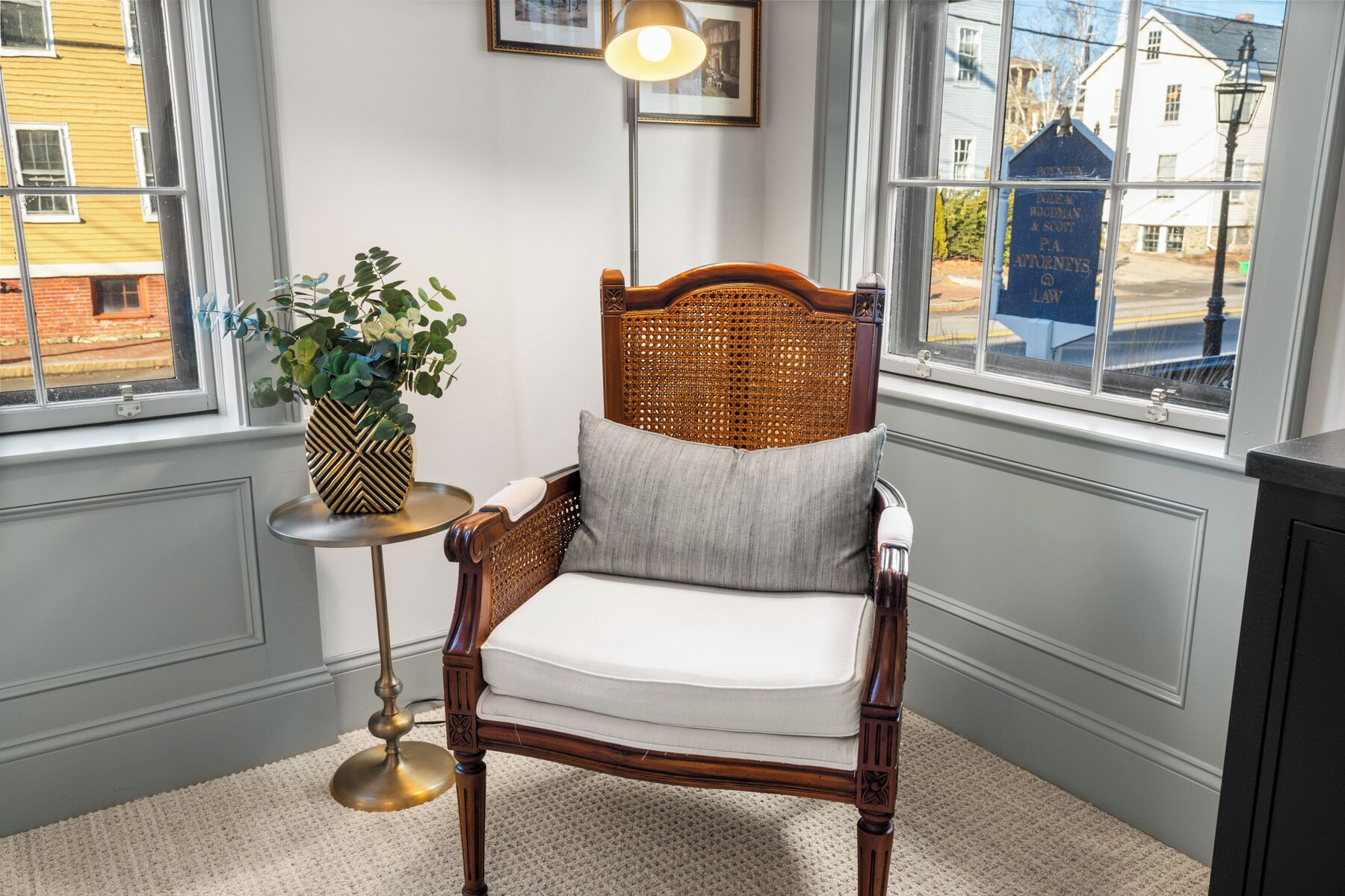 A corner room with a wooden chair, a small table with a lamp, and a view of the street through a window.