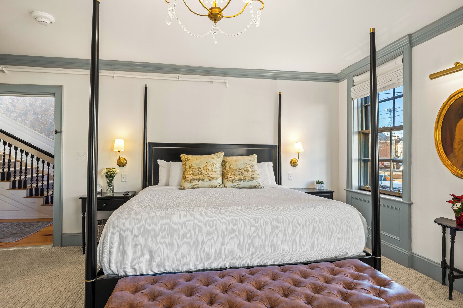 A four-poster bed with a tufted bench, a chandelier, and a view of the outside.