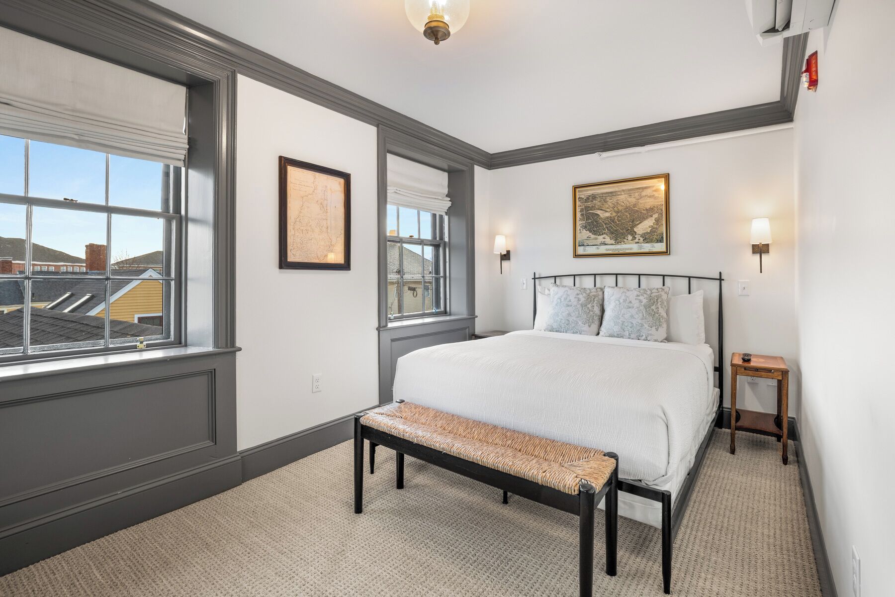 Quarto com uma cama grande, um banco, duas luminárias na parede e uma janela com vista para edifícios.