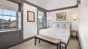 A bedroom with a large bed, a bench, two wall-mounted lamps, and a window with a view of buildings.