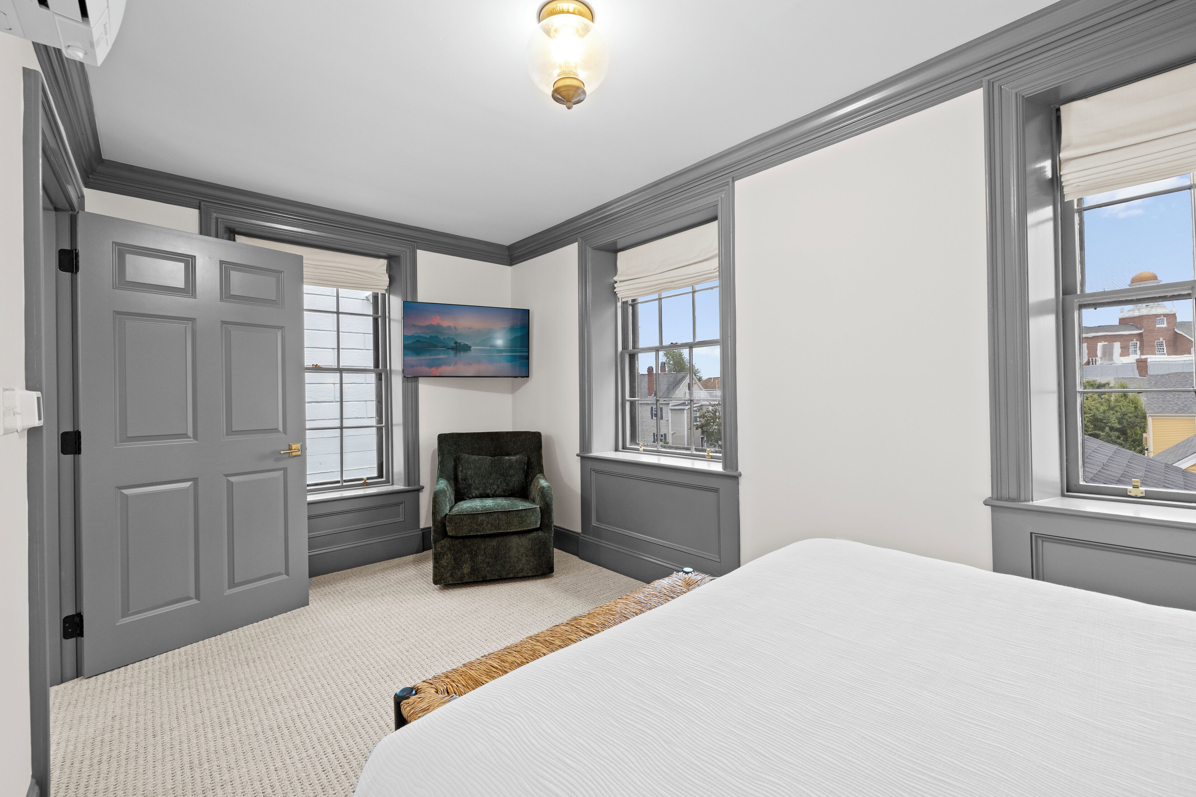 A bedroom with a large bed, a chair, a painting, and a window with a view of buildings.
