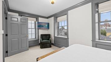 A bedroom with a large bed, a chair, a painting, and a window with a view of buildings.