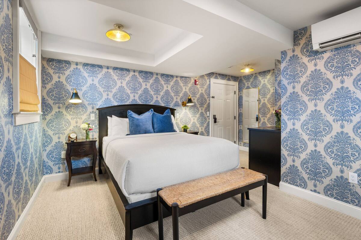 Un dormitorio con una cama grande, dos mesitas de noche, un banco, papel tapiz estampado y una lámpara en el techo.