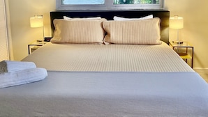 A hotel room with a bed, two bedside lamps, a window with a view of palm trees, and a painting on the wall.