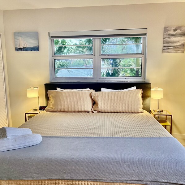 A hotel room with a bed, two bedside lamps, a window with a view of palm trees, and a painting on the wall.