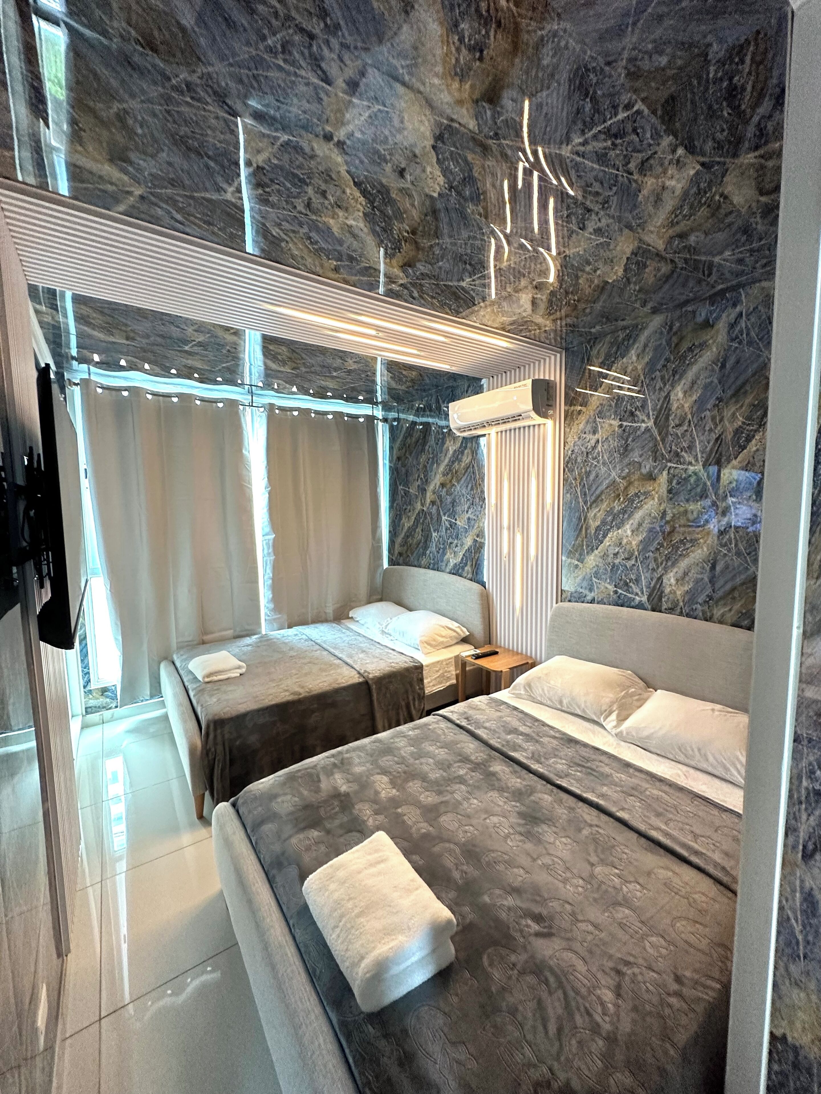 A modern hotel room with a marble wall, two beds, a nightstand, and a ceiling light fixture.