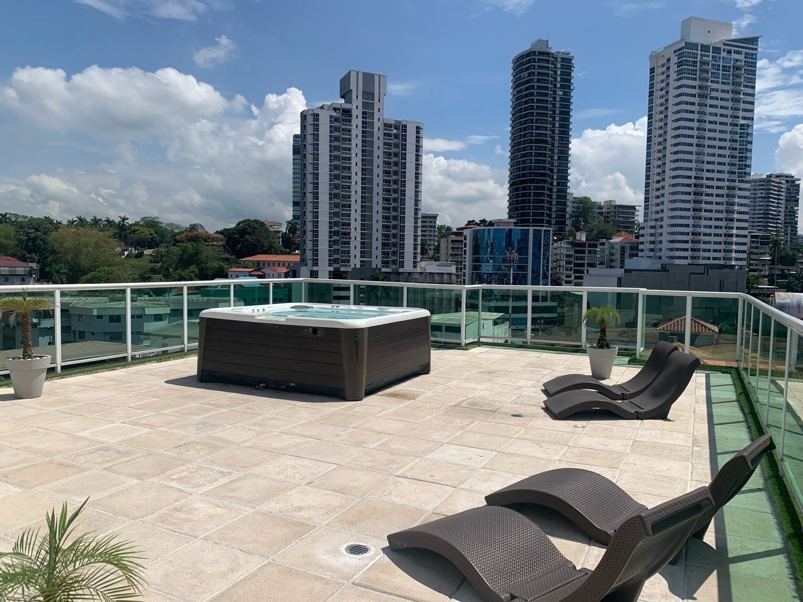 A rooftop terrace with a hot tub, lounge chairs, and a glass railing overlooking a cityscape.
