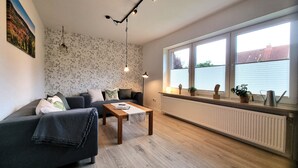 A living room with a sofa, coffee table, and a radiator. There are windows with blinds and a view of a building outside. - Heidezauber: Modern vacation apartment with terrace and fireplace (Uelzen)