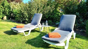 Two lounge chairs with cushions, a small table with a water bottle and glasses, and a garden with green grass and bushes. - Heidezauber: Modern vacation apartment with terrace and fireplace (Uelzen)