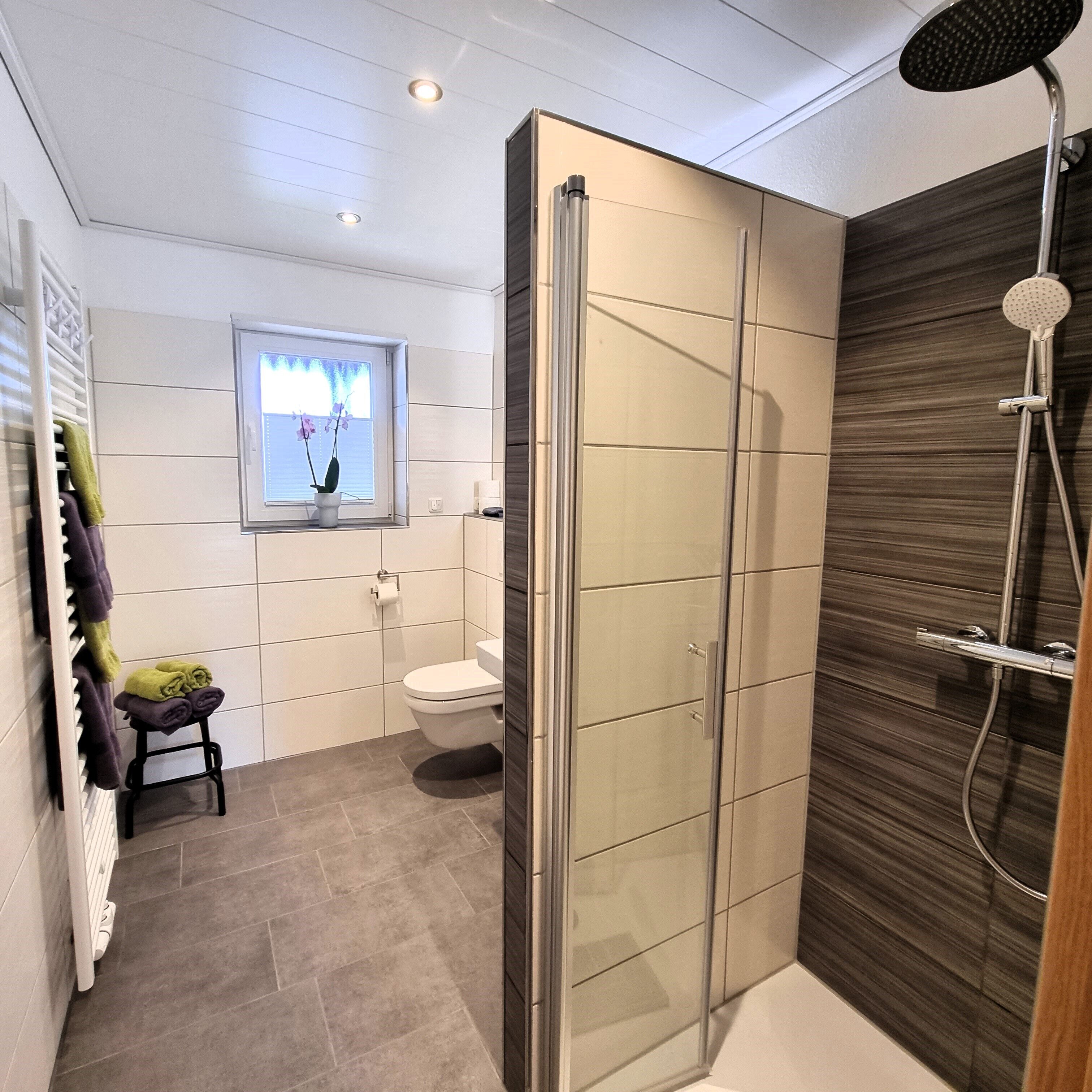 Une salle de bain moderne avec une douche à parois de verre, des toilettes et un petit tabouret sur lequel sont posées des serviettes.