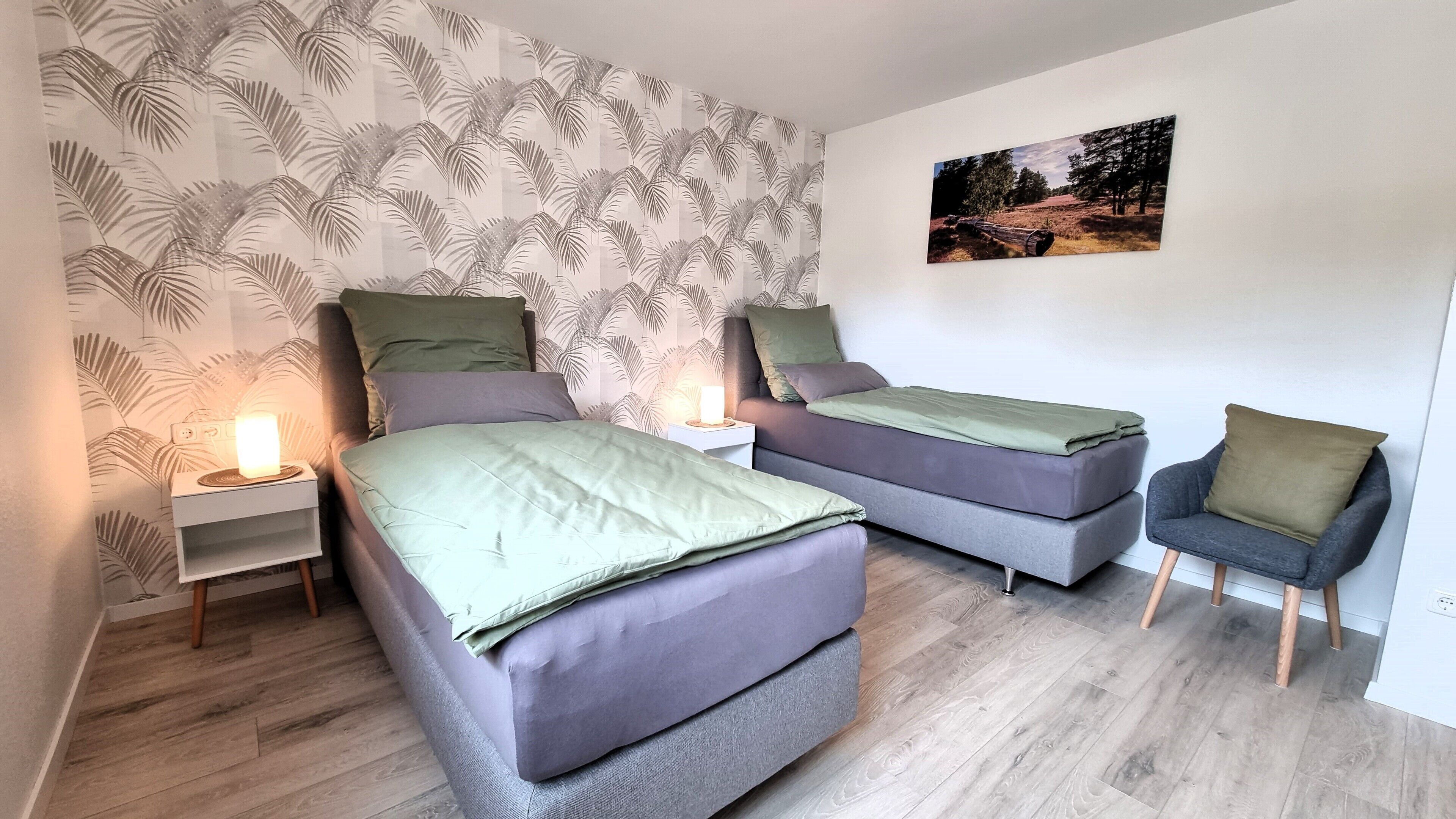 A bedroom with two beds, a nightstand with a lamp, a chair, and a wall with a tropical wallpaper and a picture.