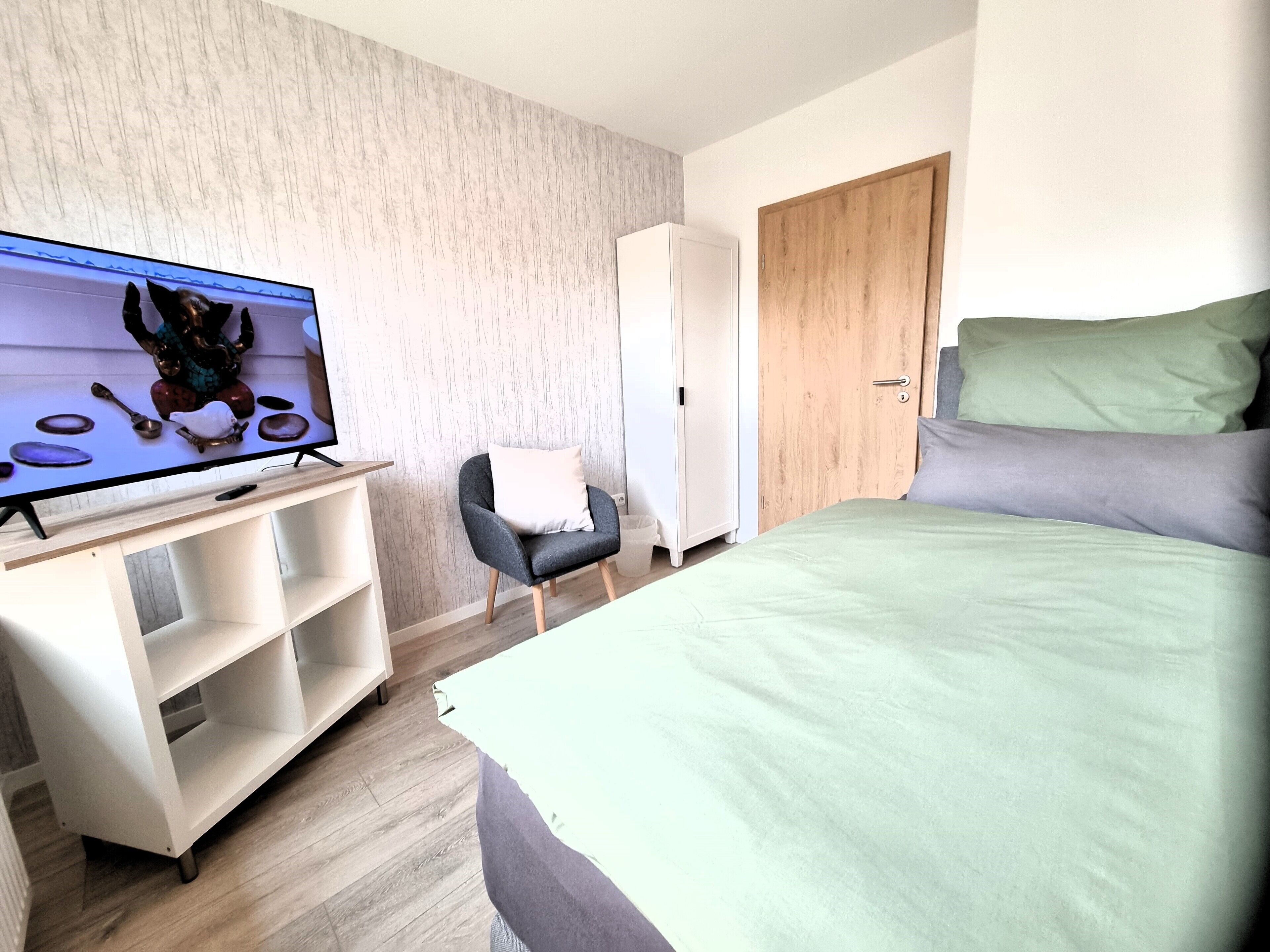 A modern hotel room with a bed, a TV on a stand, a wardrobe, and a chair.