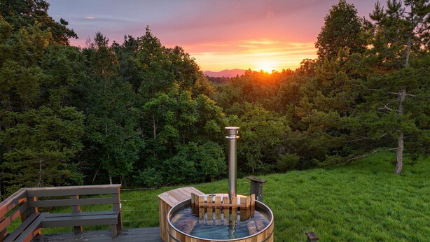 Un jacuzzi en una terraza de madera con un banco de madera y un taburete pequeño, rodeado de vegetación exuberante.