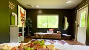 Una habitación con una cama, un sofá, un ventanal con vista a follaje, una pared con un cuadro y una bandeja con comida sobre la cama.
