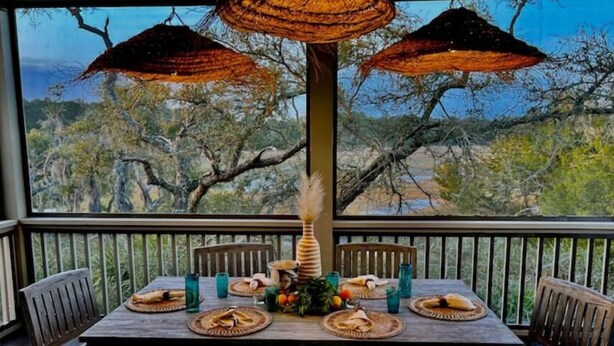 A dining table set with plates, cutlery, and glasses, surrounded by chairs, with a view of trees and a body of water through large windows. - Marshfront Villa In The Trees - Near Beach & Bay
 (Edisto Island)