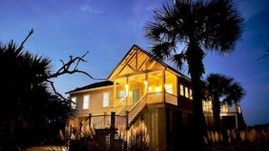 A two-story house with a balcony and a porch, surrounded by palm trees. - Marshfront Villa In The Trees - Near Beach & Bay
 (Edisto Island)