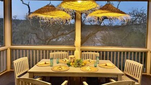 A dining area with a large table set for six, surrounded by wooden chairs. There are three hanging woven lamps above the table, and the room has large windows offering a view of trees and a clear sky. - Marshfront Villa In The Trees - Near Beach & Bay
 (Edisto Island)