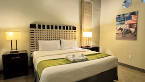A hotel room with a bed, two bedside tables, a headboard, a wall-mounted lamp, a painting, and a carpeted floor.