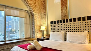 Une chambre d’hôtel avec un grand lit, un plaid rouge et une vue sur le paysage urbain, visible à travers une grande fenêtre.