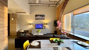 A modern living room with a sofa, a dining table set for two, a flat-screen TV, and a large window offering a view of the outdoors.