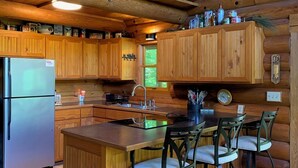 A wooden cabin kitchen with a refrigerator, wooden cabinets, a sink, and a bar area with stools. - Make Memories - Secluded Log Cabin on Marion Lake Channel with Private Dock (Richville)