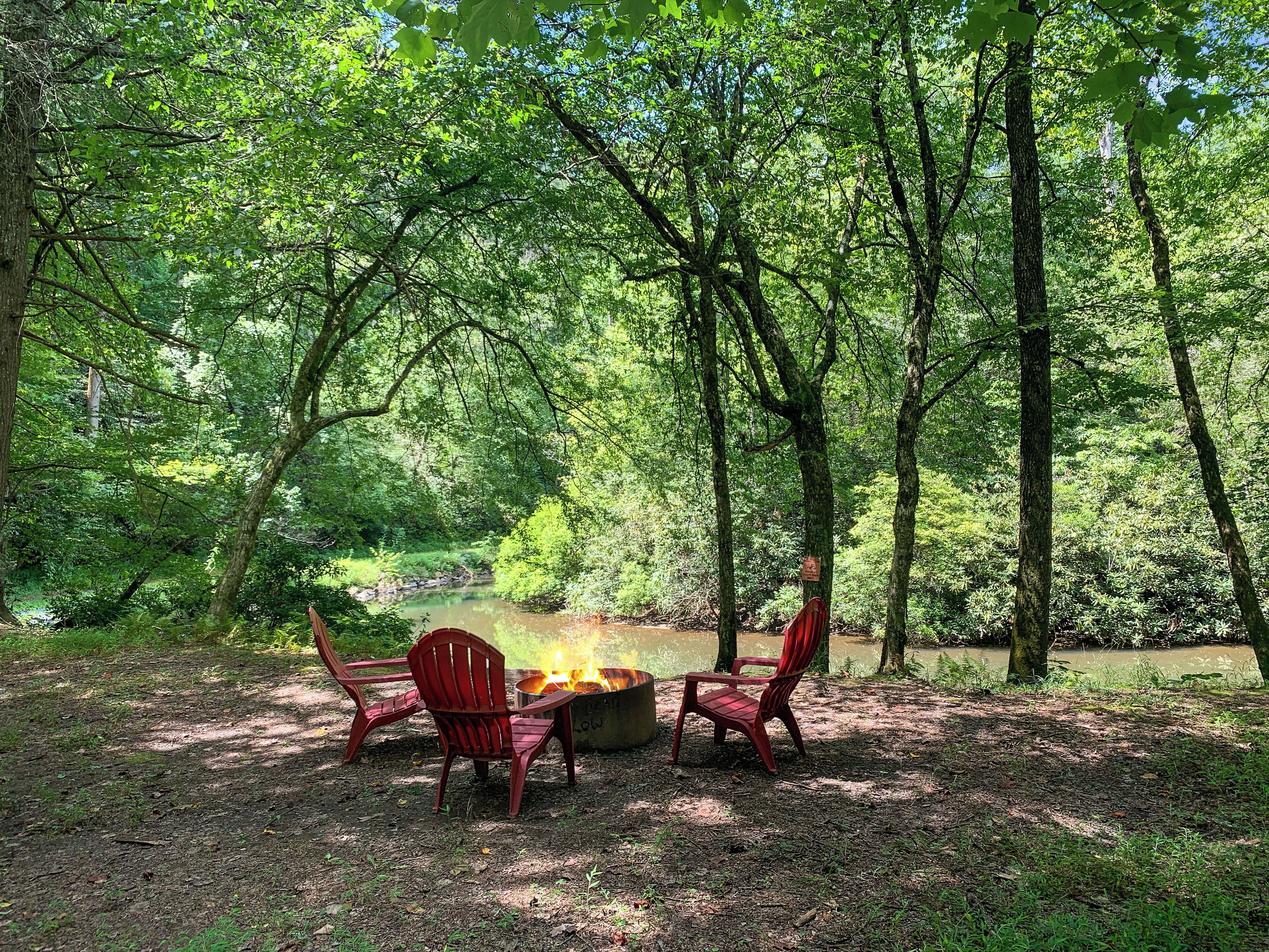 A campsite with a fire pit surrounded by red chairs and a table, set in a forested area with a pond in the background.