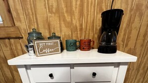 A white wooden coffee station with a sign reading "But first Coffee," a green mug, an orange mug, and a black coffee maker. - Cozy little cabin in a small town  (Caney)