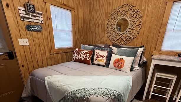 A cozy room with wooden walls, a bed with a gray bedspread, and a small table with a clock. - Cozy little cabin in a small town  (Caney)