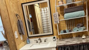 A bathroom with a wooden wall, a white vanity with a mirror, a towel rack with towels, and a shelf with toiletries. - Cozy little cabin in a small town  (Caney)