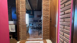 Un couloir avec un mur de briques d’un côté et un mur rose de l’autre, qui mène à un espace cuisine comprenant une table à manger et des chaises.