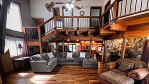 A spacious living area with a wooden floor, a large sectional sofa, a smaller sofa, and a patterned ottoman. A staircase leads to an upper level with a balcony. - Family Cabin in Graeagle CA w/King Bed & EV Charge (Graeagle)