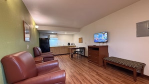 A living room with a sofa, a bench, a television, and a kitchen in the background. - Relax Inn & Suites (Mattoon)