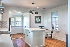 Una cocina con suelo de madera, muebles blancos, una isla central, iluminación colgante y vistas al exterior.