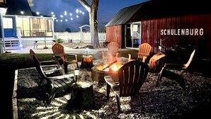Een terras met Adirondack-stoelen en een vuurkorf, omgeven door een houten terras en een rode schuur met de tekst 