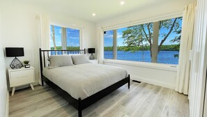 Ein Schlafzimmer mit einem Bett, Nachttischen und großen Fenstern mit Blick auf einen See und Bäume.