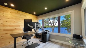 Ein zeitgemäßer Arbeitsbereich mit großem Schreibtisch, ergonomischem Stuhl und zwei Bildschirmen. Er verfügt über eine Holzvertäfelung, ein großes Fenster mit Seeblick und einen Glastisch.