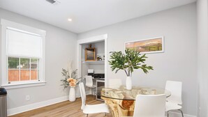 A modern dining area with a glass table, white chairs, a wooden accent wall, a view of the outdoors, and a shelf with decorative items.