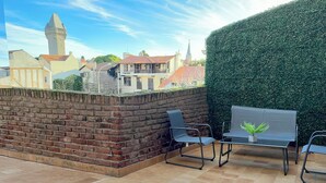 Eine Dachterrasse mit Backsteinmauer, zwei Stühlen, einer Bank und Blick auf Gebäude sowie einen Turm im Hintergrund.