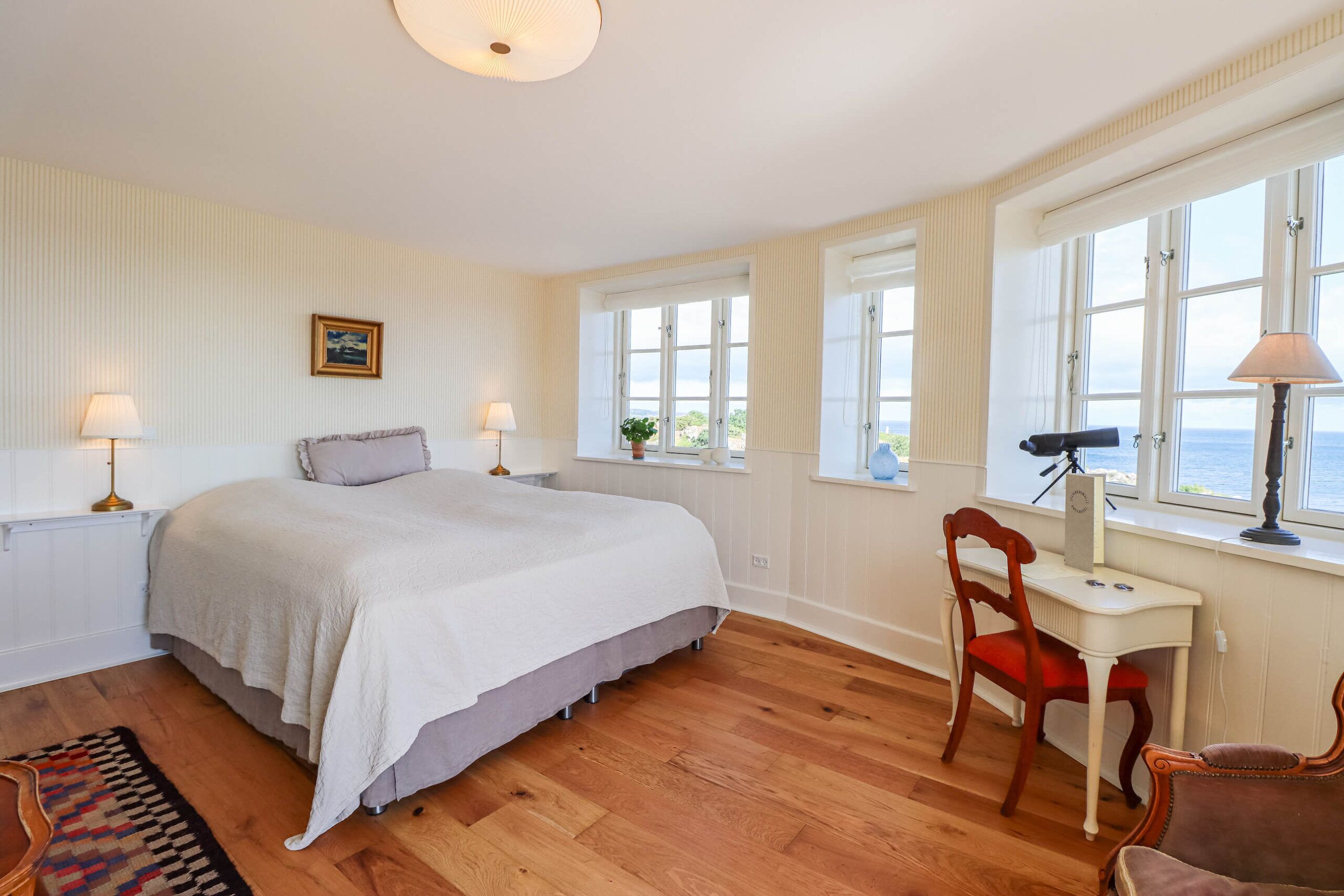 A bedroom with a large bed, a desk with a chair, and a view of the sea through the windows.