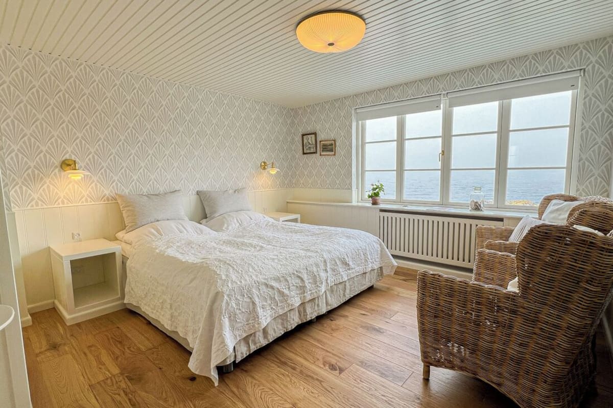 A bedroom with a bed, nightstands, a wicker chair, a window with a view of the sea, and patterned wallpaper.