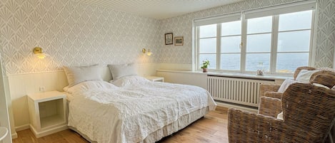 Un dormitorio con cama, mesitas de noche, una silla de mimbre, una ventana con vista al mar y papel tapiz estampado.