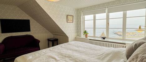 A bedroom with a large bed, a red sofa, a window with a view of the sea, and a decorative ceiling light.