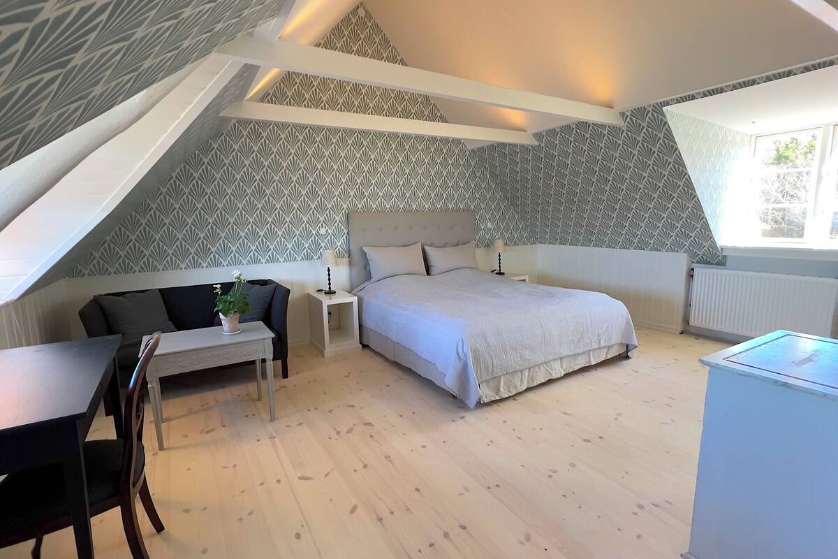 A bedroom with a sloped ceiling, a bed with a headboard, a desk with a chair, a small table with a plant, and a radiator.
