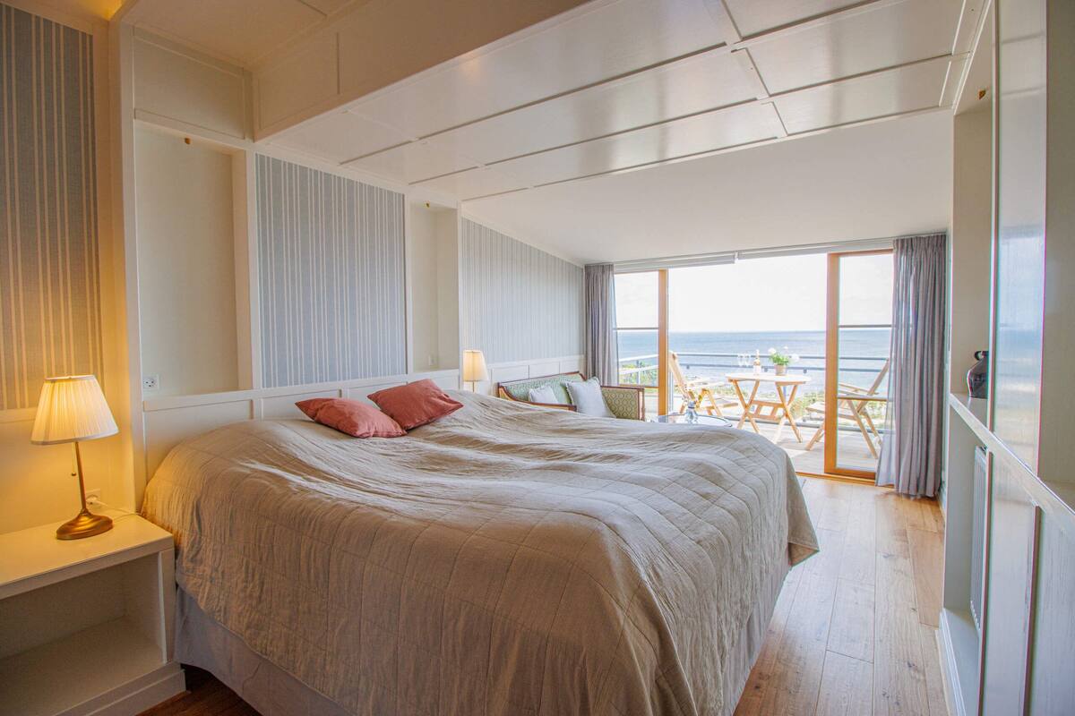 A spacious bedroom with a large bed, a nightstand with a lamp, and a view of the sea through a balcony.