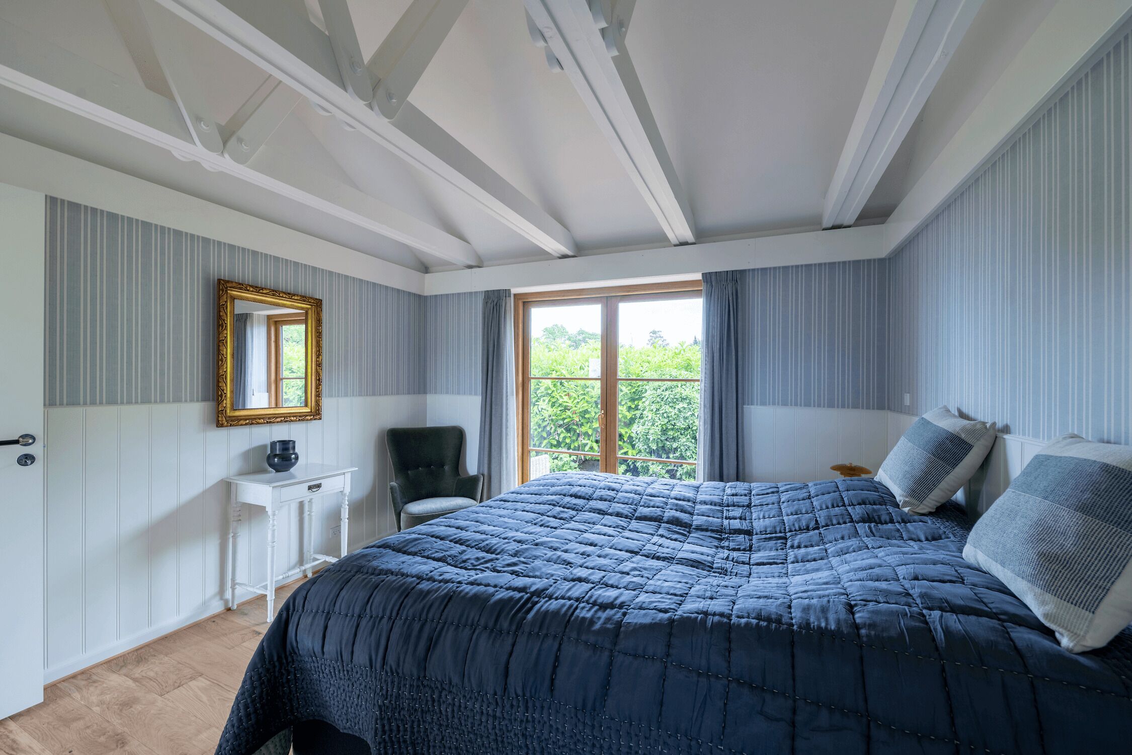 A bedroom with a large bed, a bedside table, a chair, a window with curtains, and a mirror on the wall.